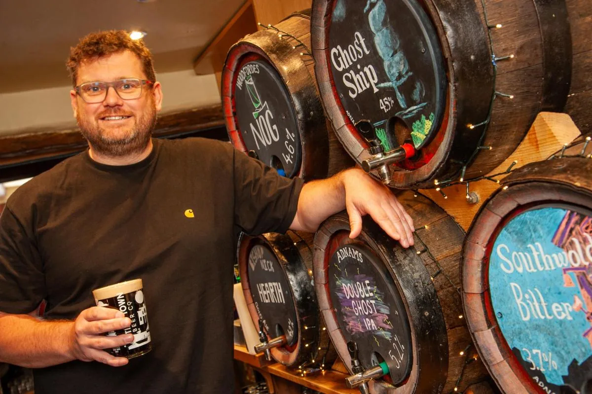Tom West smiling behind the bar, holding a Tiny Town pint, next to casks of real ale